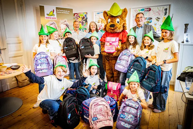 Zum Auftakt der jährlichen Schulranzenaktion des Familienfonds ROBIN GOOD übergab Maskottchen Robbie heute unter den Kunstwerken der „Starken Pänz“ symbolisch Ranzen an die Botschafterkinder von der OGS Servatius und der OGS Donatus. In der Fachstelle für Suchtprävention update von Caritas und Diakonie nahmen die Schüler*innen die Schultaschen stellvertretend für sozial benachteiligte Kinder entgegen.  | Foto: Robin Good