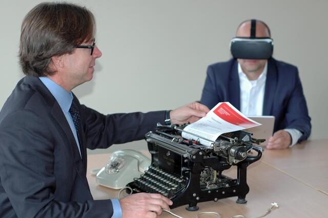 Die Digitale Transformation der Arbeitswelt, festgehalten auf einem Foto: Links Johannes Klapper mit „Wählscheibentelefon“, mechanischer Schreibmaschine und Papier. Im Hintergrund Thorsten Rolfsmeier mit VR-Brille und Tablet in der virtuellen Welt unterwegs. | Foto: Cuvelier / AA
