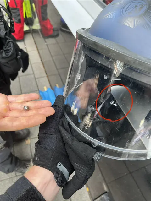 Mehrere vermummte Personen haben die Einsatzkräfte gezielt mit Stahlkugeln beschossen. Ein Beamter hält eine solche Kugel in der Hand, die durch den Beschuss entstandene Beschädigung im Visier ist rot markiert.  | Foto: Polizei Köln
